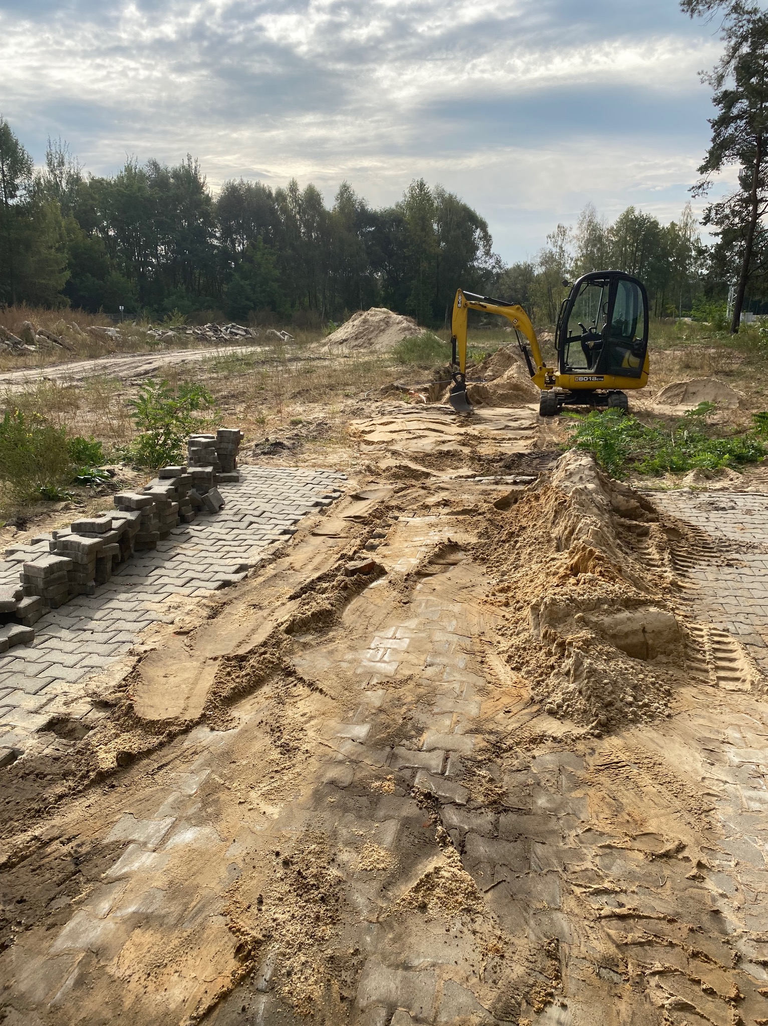 Żółta mini koparka wyrównuje teren pod budowę chodnika z szarej kostki brukowej, widoczne ślady opon i hałdy piasku, w tle las.