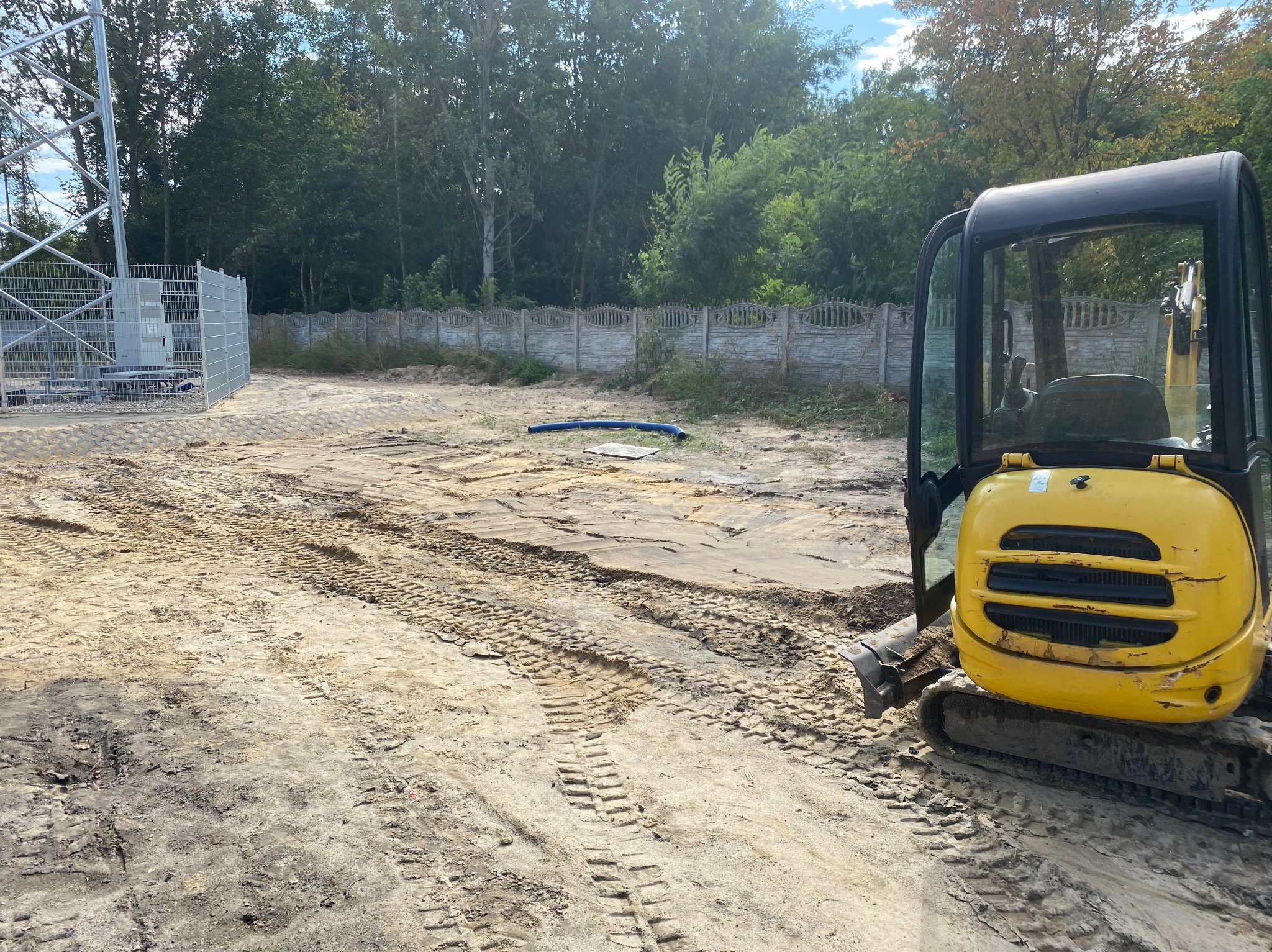 Żółta mini-koparka na placu budowy, widoczne ślady opon na piasku, w tle ogrodzenie z betonowych elementów i metalowa konstrukcja wieży.