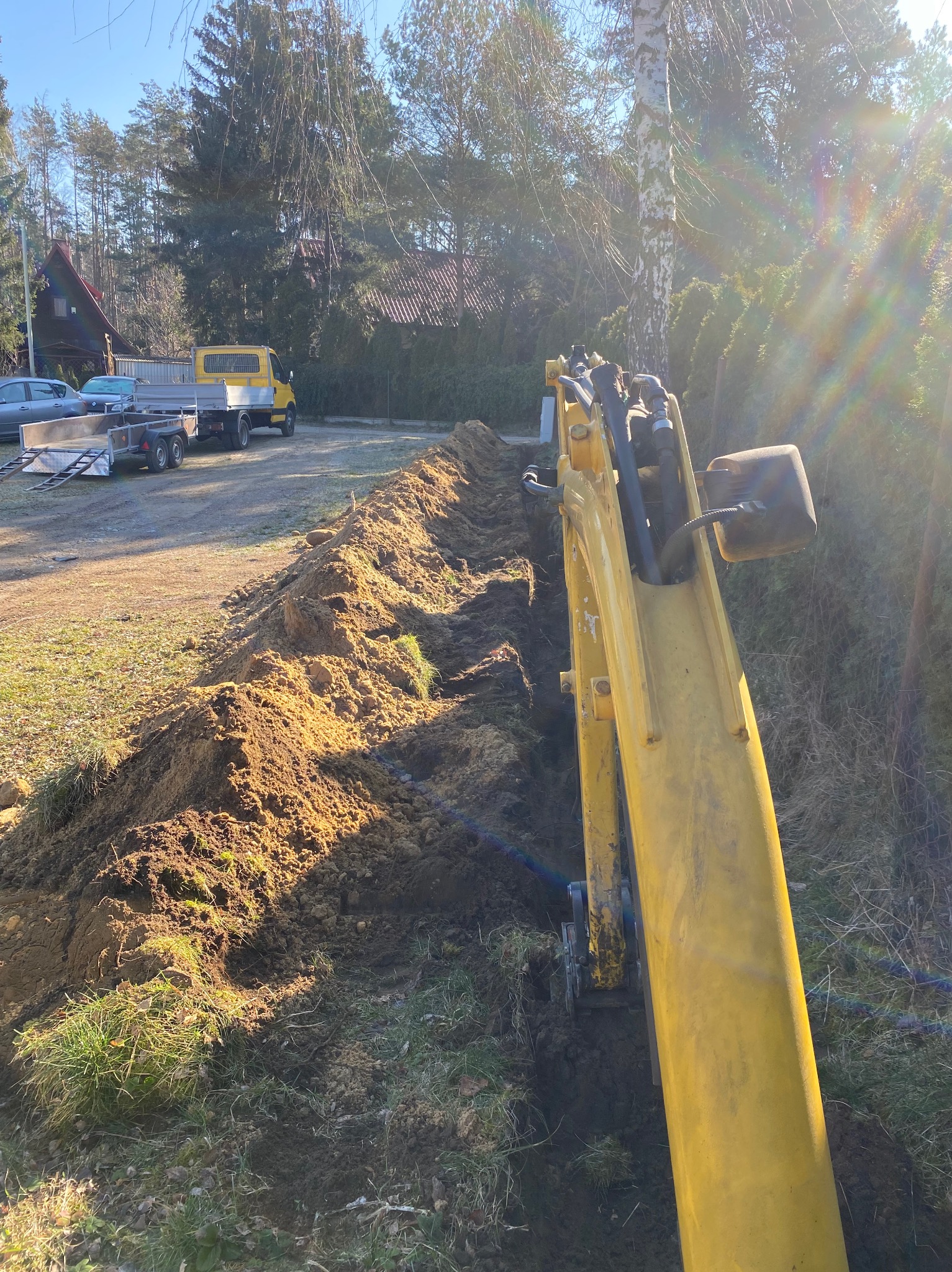 Żółta koparka w trakcie robót ziemnych; wykopany wąski rów w gruncie, pryzma ziemi obok, w tle żółta ciężarówka z przyczepą na posesji.