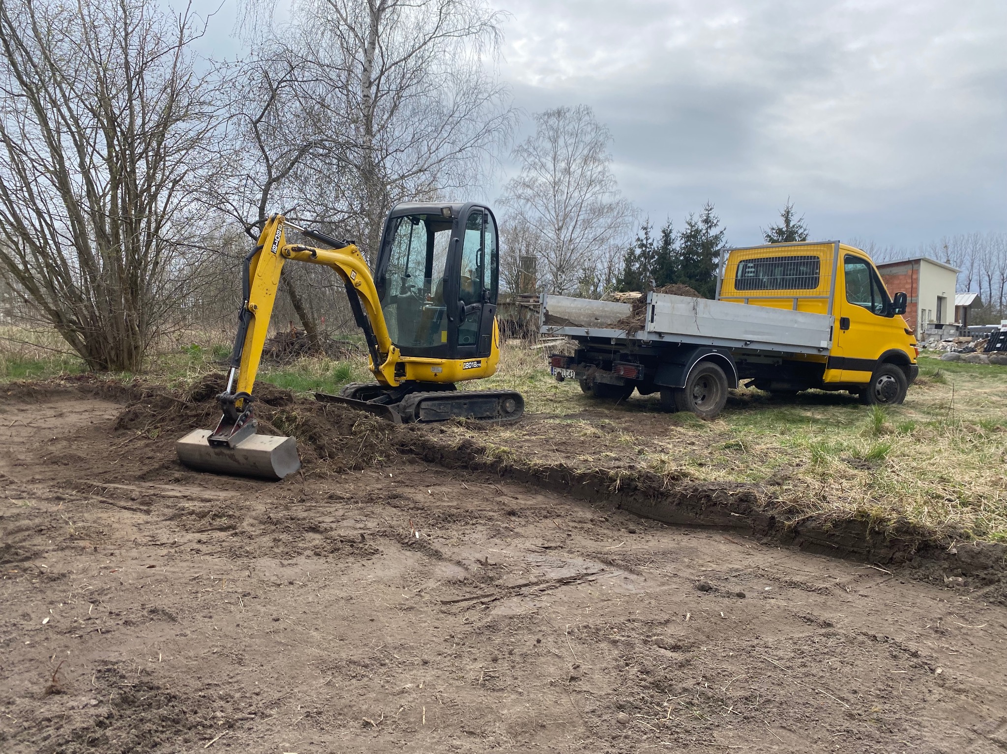 Żółta mini koparka z łyżką i żółta ciężarówka na działce z wykopanym rowem w ziemi, przygotowanie terenu pod przyszłą instalację.