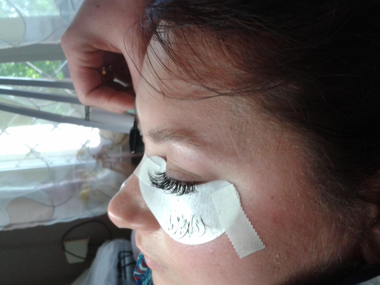 Zbliżenie na oko kobiety podczas zabiegu przedłużania rzęs, widoczna ochronna naklejka pod okiem i dłoń osoby wykonującej zabieg.