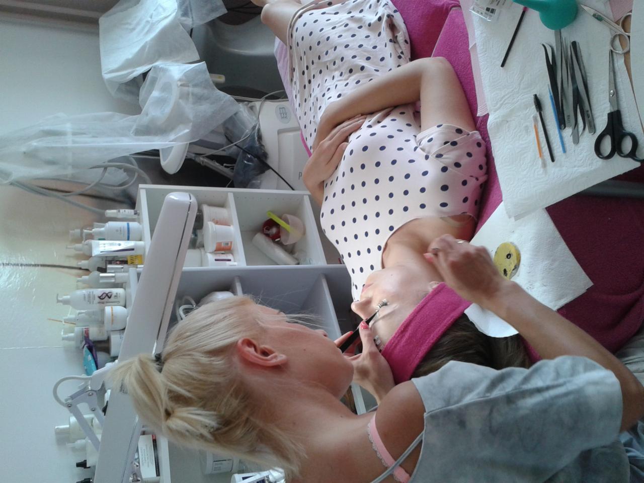 Klientka leży na różowym łóżku podczas zabiegu przedłużania rzęs, kosmetyczka aplikuje rzęsy pęsetą, obok leżą narzędzia na papierowym ręczniku, w tle widoczne kosmetyki i lampa powiększająca.