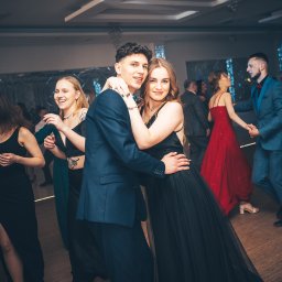 Jakub Chlebda Fotografia - Para młodych ludzi w eleganckich strojach, obejmująca się na parkiecie podczas imprezy; w tle tańczący goście i dekoracje świetlne.