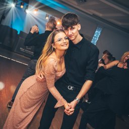 Jakub Chlebda Fotografia - Para młodych trzymająca się za ręce podczas pierwszego tańca na sali balowej z parkietem i oświetleniem scenicznym, widoczni goście w tle.