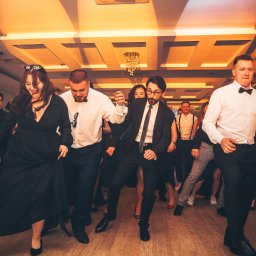 Jakub Chlebda Fotografia - Grupa elegancko ubranych osób tańczy na parkiecie pod dekoracyjnym sufitem z żyrandolami, widoczne dynamiczne ruchy i radosna atmosfera.