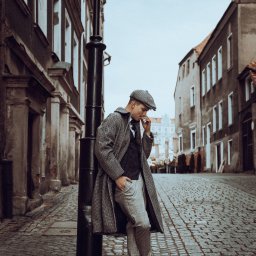 Jakub Chlebda Fotografia - Mężczyzna w tweedowym płaszczu i kaszkiecie opiera się o czarną latarnię na brukowanej ulicy, w tle kamienice.
