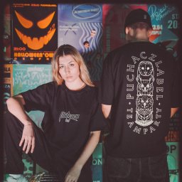 Jakub Chlebda Fotografia - Dwoje młodych ludzi pozuje w ciemnych ubraniach na tle plakatów imprezowych, ona siedzi, on stoi tyłem, prezentując nadruk z sowami na koszulce.