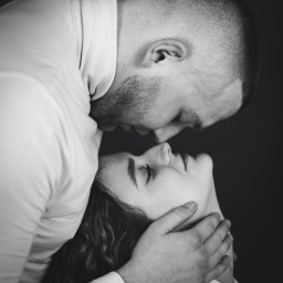 Jakub Chlebda Fotografia - Czarno-białe zbliżenie pary: mężczyzna z krótkimi włosami obejmuje kobietę, delikatnie dotykając jej szyi dłonią; oboje mają zamknięte oczy.
