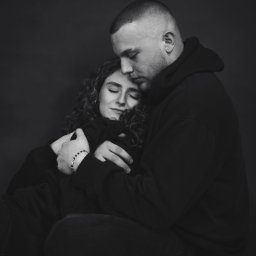 Jakub Chlebda Fotografia - Czarno-białe zdjęcie objętej pary w bluzach z kapturami, kobieta z zamkniętymi oczami i nosem z kolczykiem, mężczyzna z krótkimi włosami przytula ją od tyłu, oboje siedzą na podłodze.