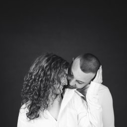 Jakub Chlebda Fotografia - Czarno-białe zbliżenie na parę w białych koszulach, dotykających się czołami, z ciemnym tłem.