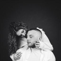 Jakub Chlebda Fotografia - Czarno-białe portretowe ujęcie pary w białych koszulach na ciemnym tle; kobieta z kręconymi włosami obejmuje mężczyznę, dotykając jego policzka.