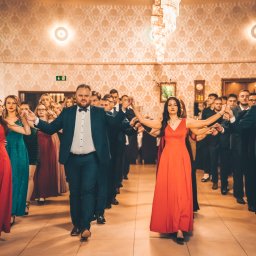 Jakub Chlebda Fotografia - Grupa elegancko ubranych osób ustawiona w rzędach, tańcząca poloneza na sali balowej z żyrandolem i wzorzystą tapetą.