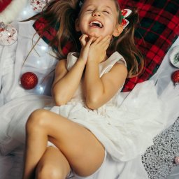 Jakub Chlebda Fotografia - Dziewczynka w białej sukience leży na pledzie w kratę z motywem świątecznym, trzymając dłonie przy twarzy i szeroko się uśmiechając, z dekoracjami świątecznymi wokół.