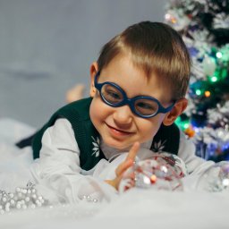 Jakub Chlebda Fotografia - Uśmiechnięty chłopiec w okularach leży na białym tle z choinką w tle, trzymając przezroczystą bombkę z czerwonymi wzorami.