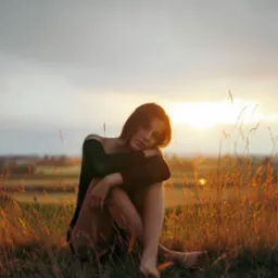 Młoda kobieta w swetrze z odkrytym ramieniem, siedząca w trawie o zachodzie słońca, opierająca głowę na kolanach.