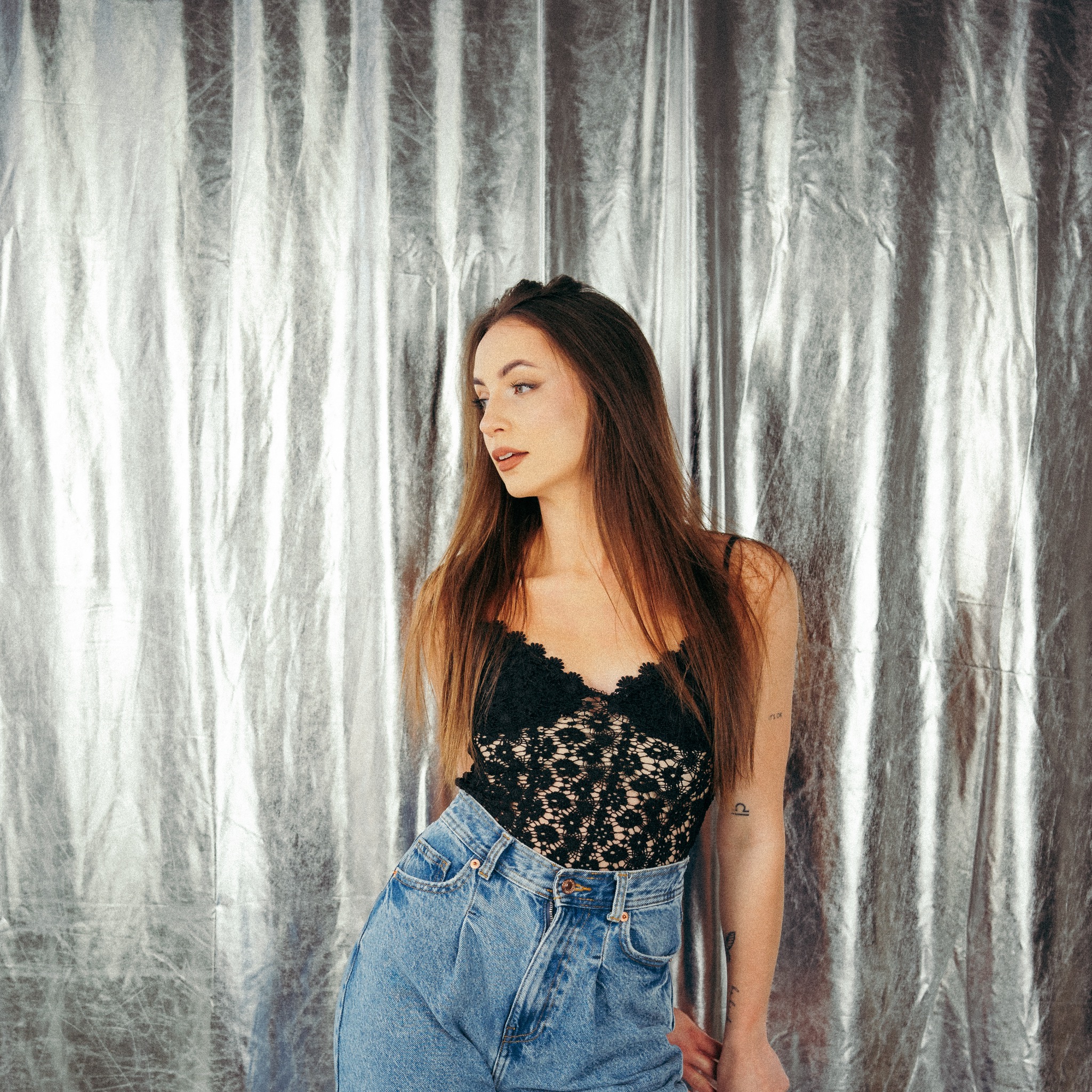 Kobieta w czarnej koronkowej bluzce i dżinsach pozuje na tle błyszczącej srebrnej tkaniny, odwrócona w lewo, z widocznym tatuażem na ramieniu.