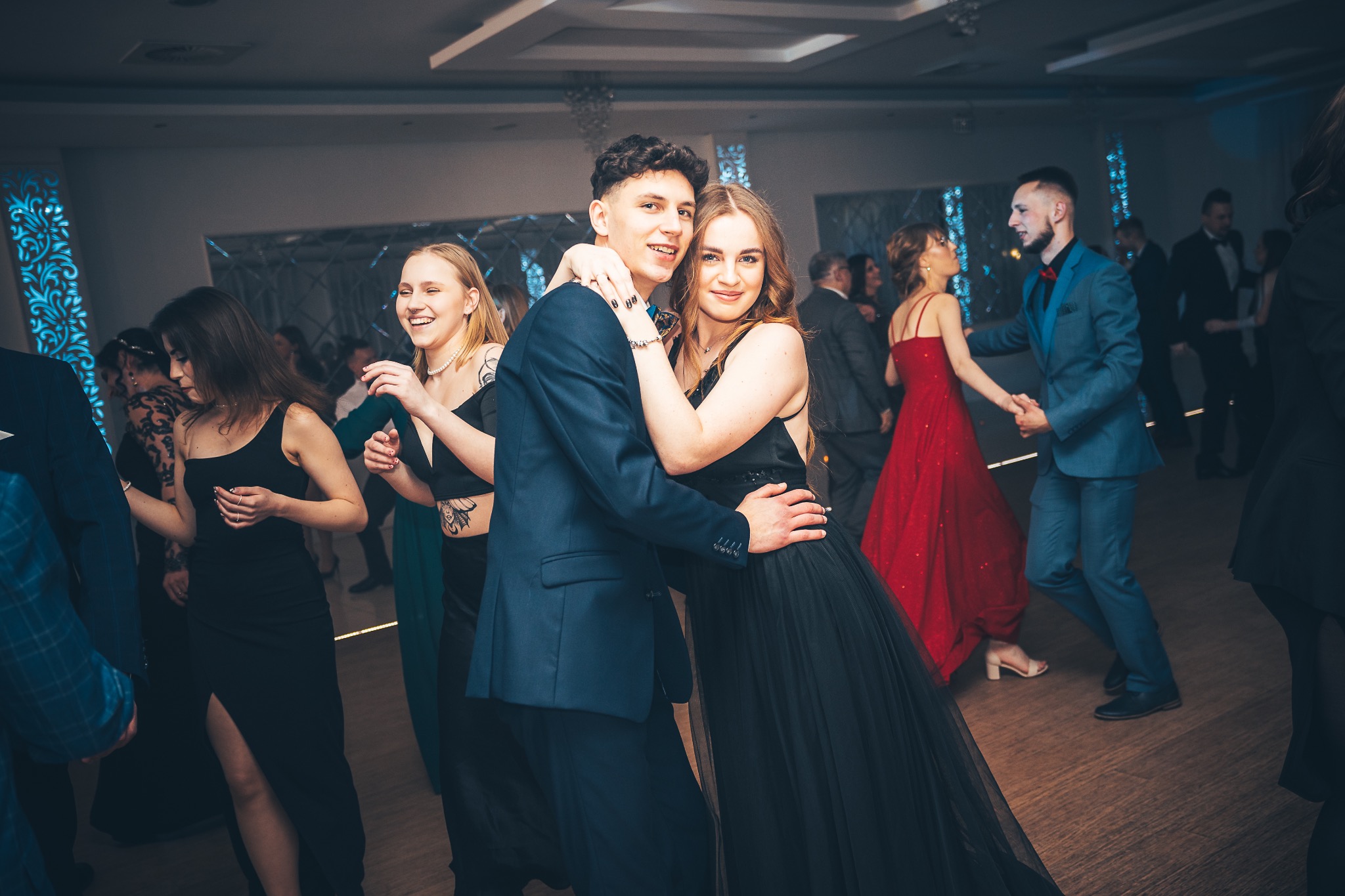Para młodych ludzi w eleganckich strojach, obejmująca się na parkiecie podczas imprezy; w tle tańczący goście i dekoracje świetlne.