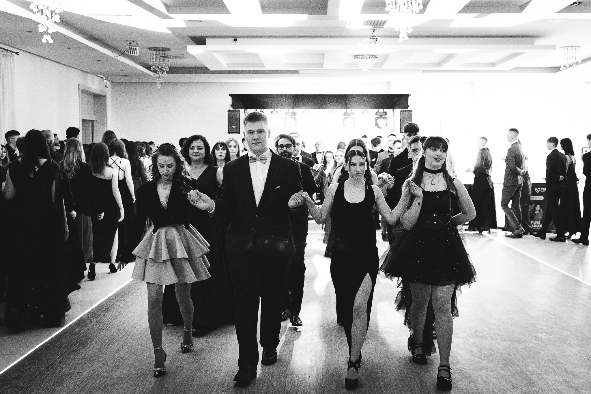 Czarno-białe zdjęcie grupy młodych ludzi, prawdopodobnie maturzystów, podczas poloneza na sali balowej z parkietem i oświetleniem.
