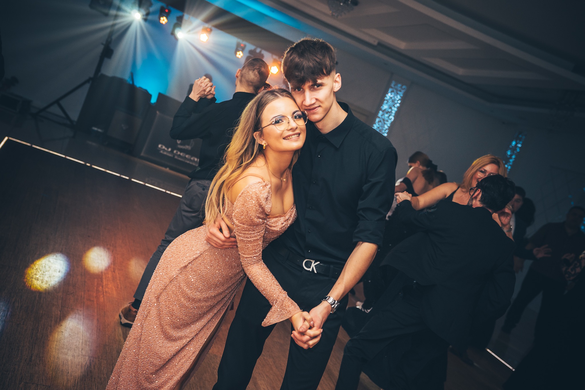 Para młodych trzymająca się za ręce podczas pierwszego tańca na sali balowej z parkietem i oświetleniem scenicznym, widoczni goście w tle.