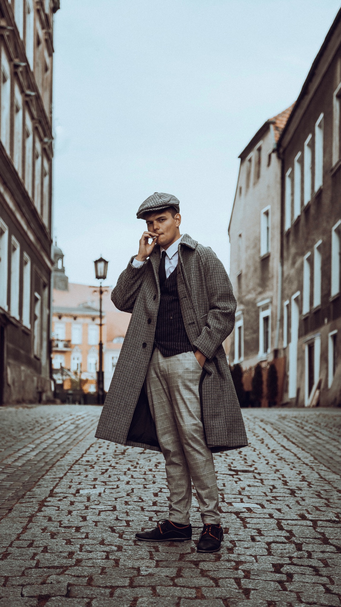 Mężczyzna w płaszczu w stylu retro i kaszkiecie pozuje z papierosem na brukowanej ulicy w Gnieźnie, otoczony starymi kamienicami.