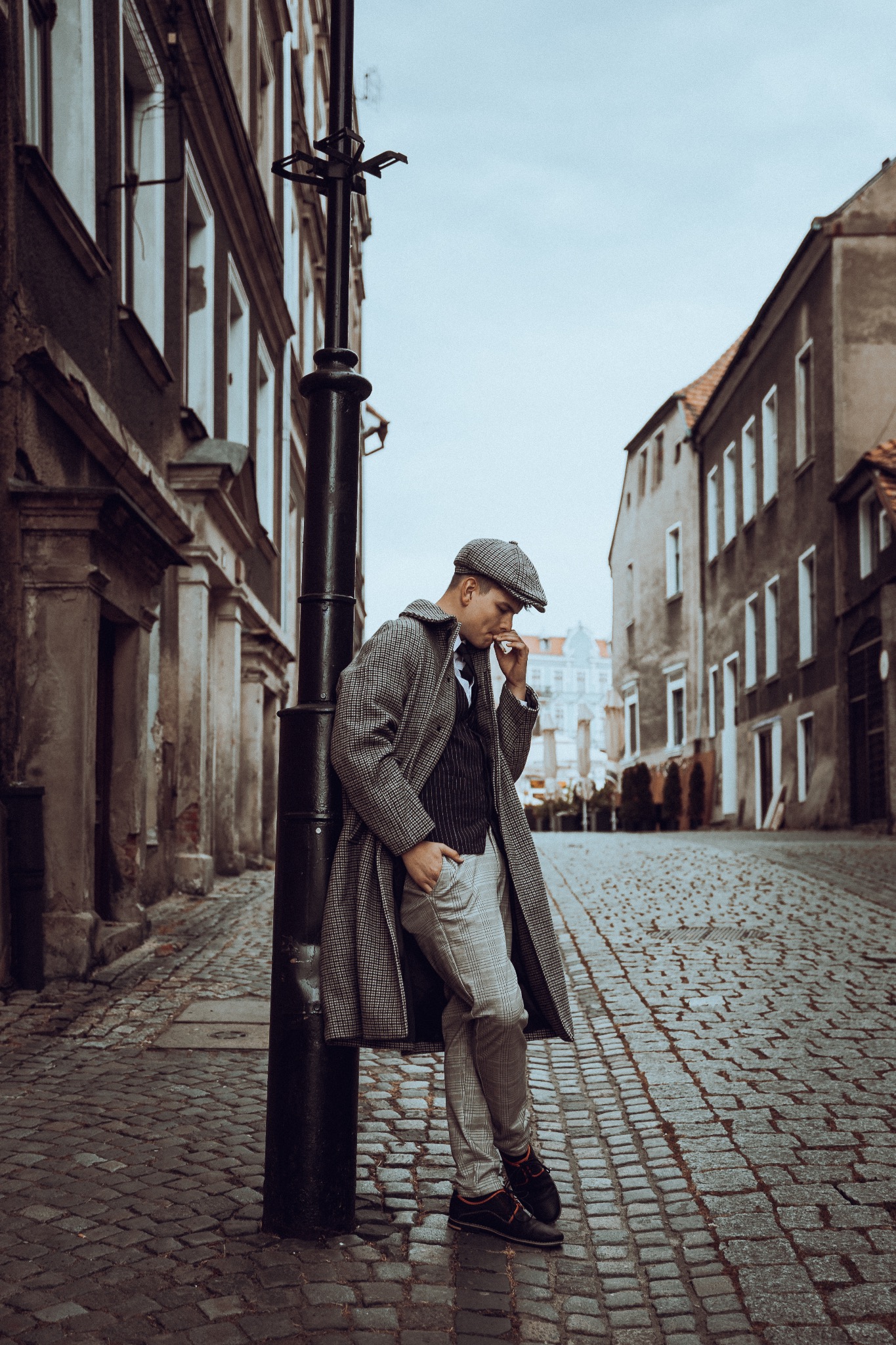 Mężczyzna w tweedowym płaszczu i kaszkiecie opiera się o czarną latarnię na brukowanej ulicy, w tle kamienice.