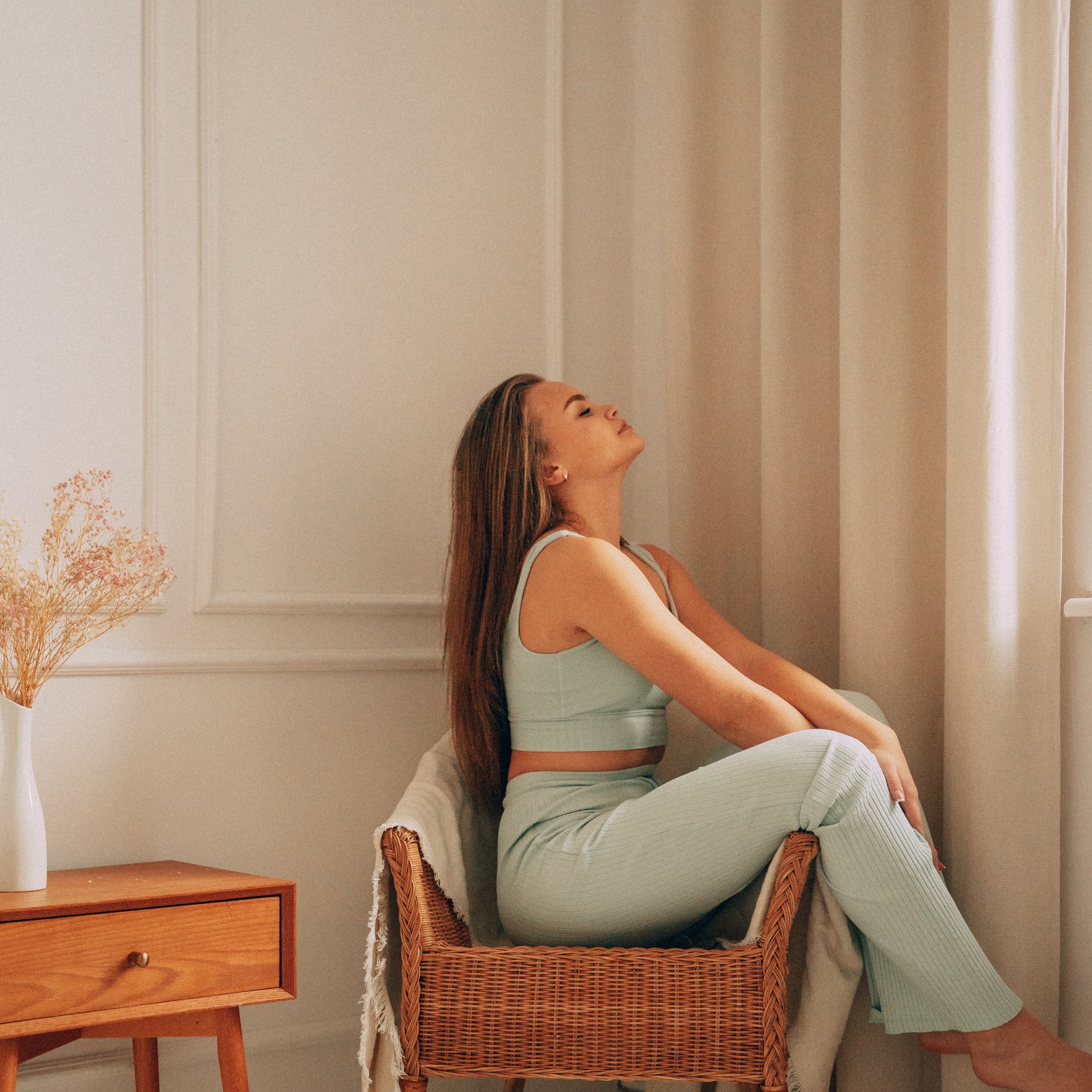 Kobieta w pastelowym, prążkowanym komplecie siedzi na wiklinowym fotelu, odchylając głowę do tyłu w jasnym, minimalistycznym wnętrzu z drewnianą szafką nocną i zasłonami.