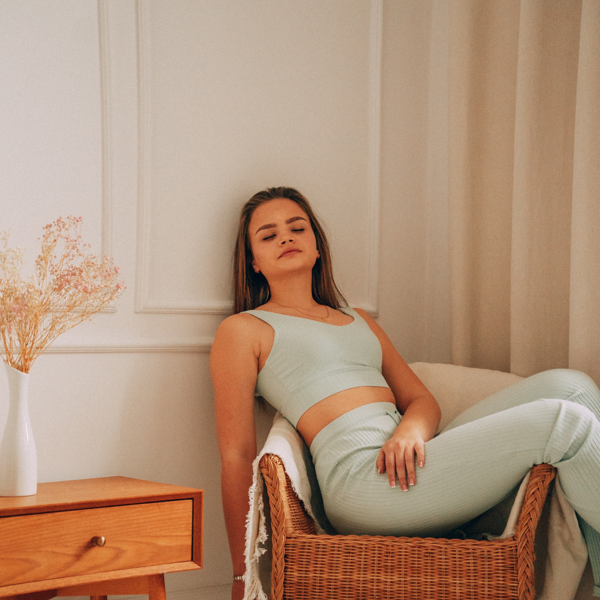 Kobieta w pastelowym komplecie siedzi w wiklinowym fotelu, opierając się o ścianę z dekoracyjną sztukaterią, obok stolik z wazonem suszonych kwiatów.