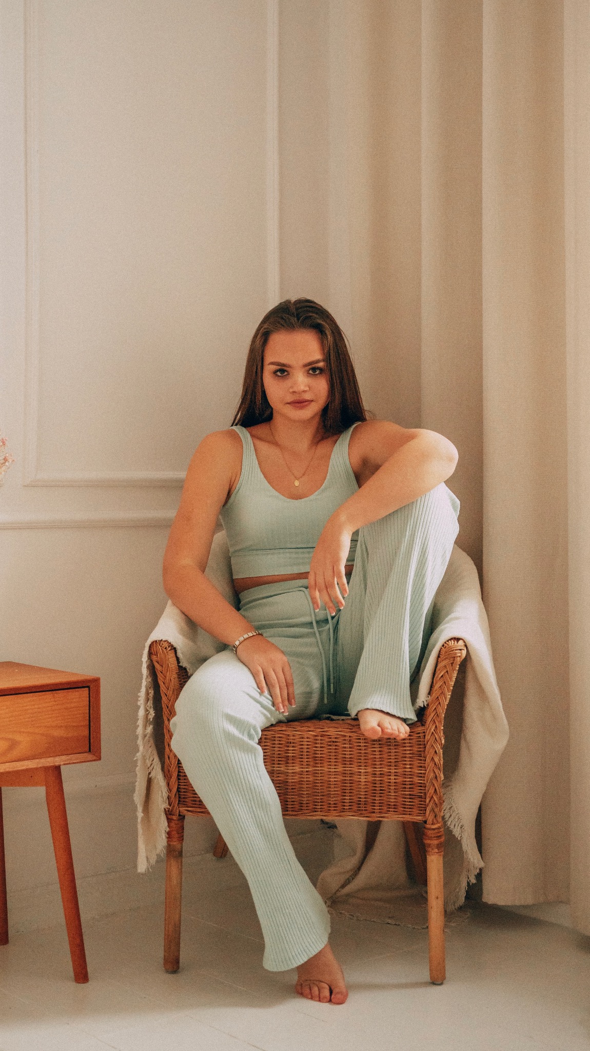 Portret kobiety w błękitnym, prążkowanym komplecie siedzącej w wiklinowym fotelu na tle jasnego wnętrza z drewnianym stolikiem nocnym.