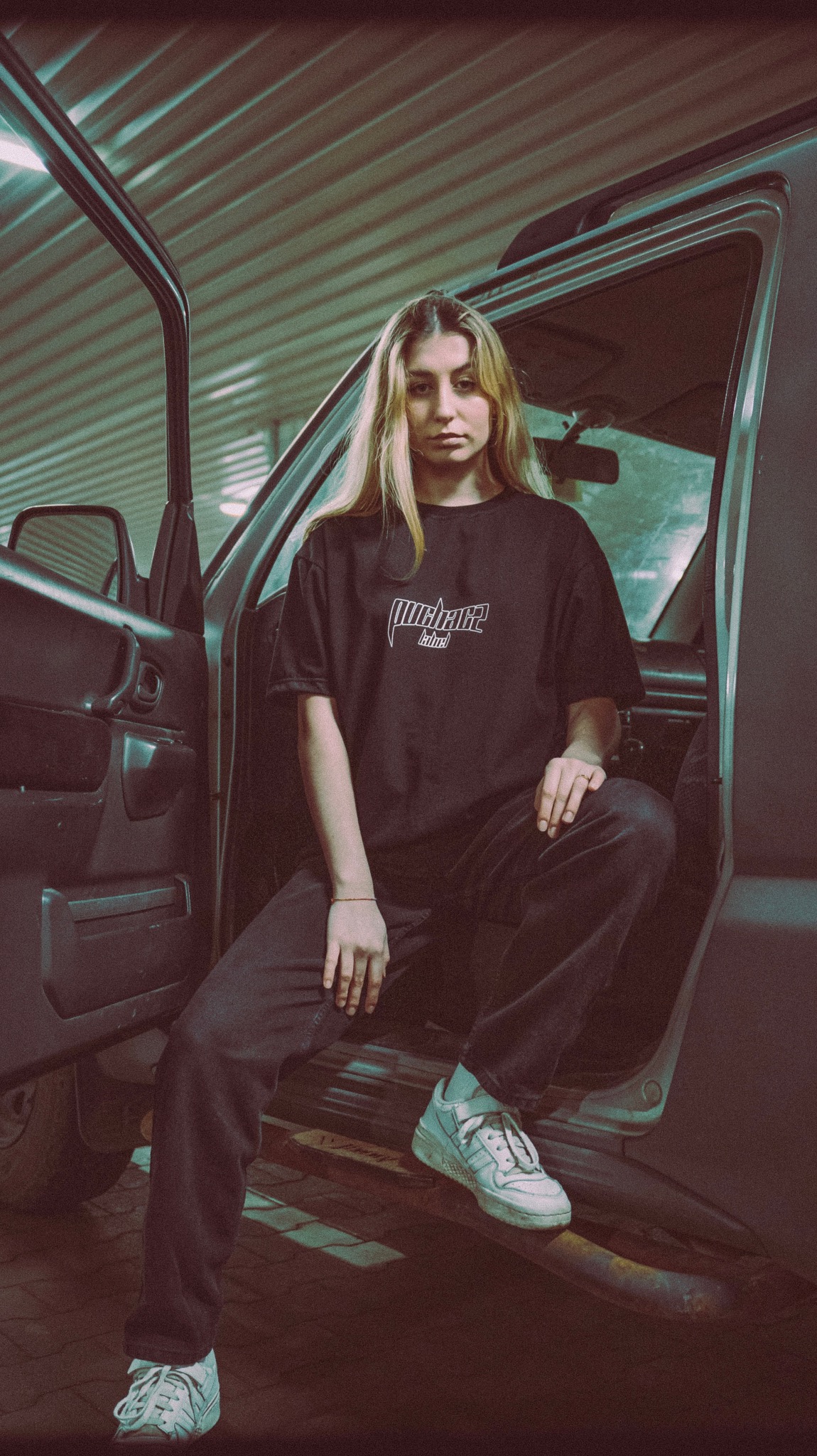 Młoda kobieta z blond włosami siedzi w samochodzie ubrana w czarny T-shirt z białym napisem, czarne spodnie i białe sportowe buty.