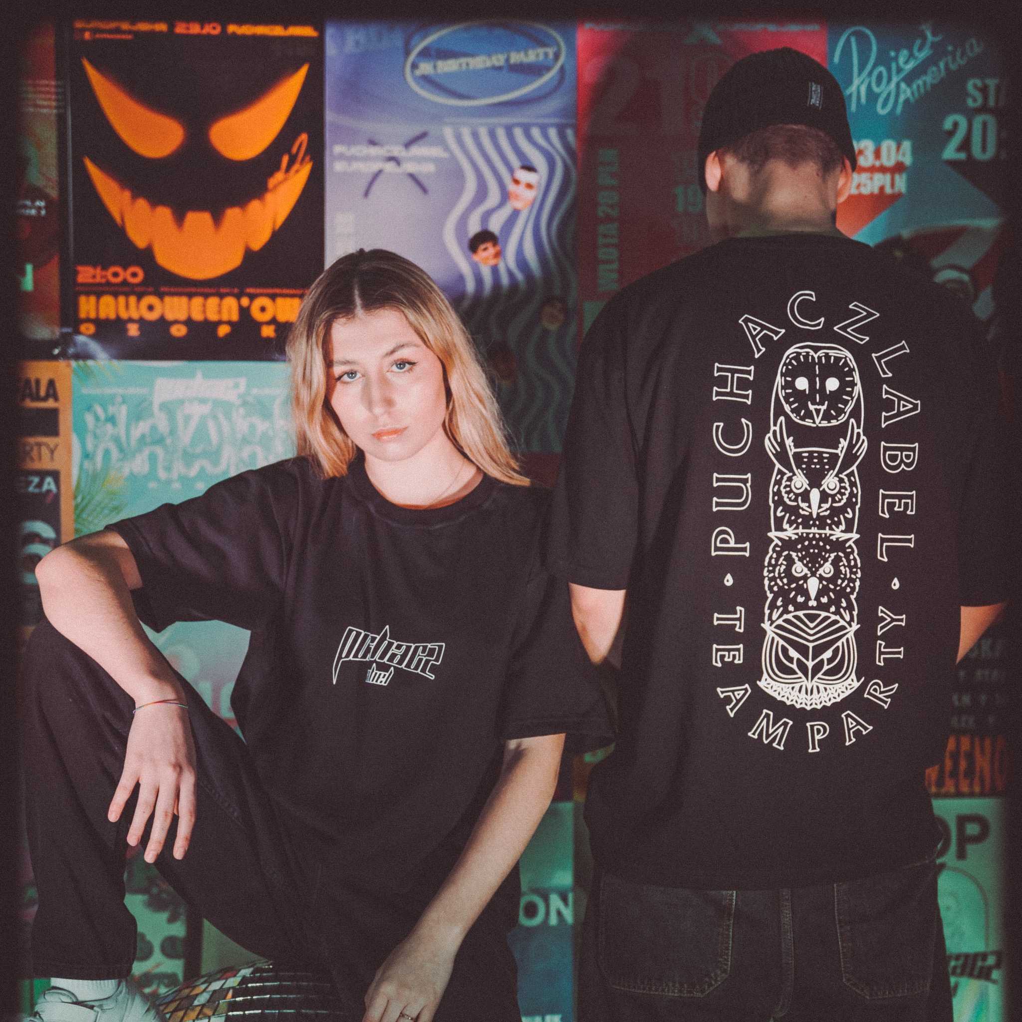 Dwoje młodych ludzi pozuje w ciemnych ubraniach na tle plakatów imprezowych, ona siedzi, on stoi tyłem, prezentując nadruk z sowami na koszulce.