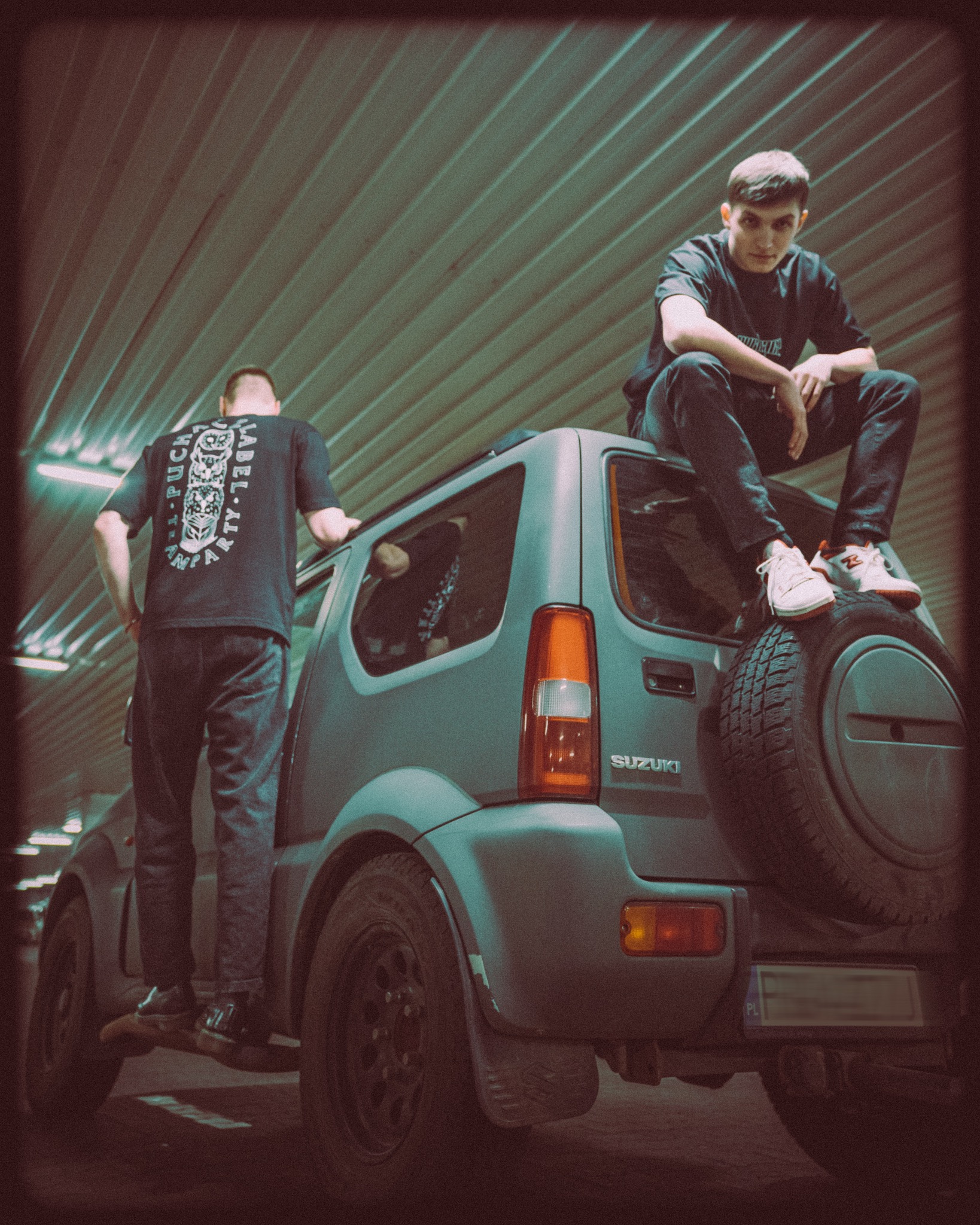 Dwóch młodych mężczyzn pozuje na i obok szarego Suzuki Jimny na parkingu podziemnym, jeden siedzi na kole zapasowym, drugi stoi opierając się o dach, obaj ubrani w czarne koszulki z nadrukami.