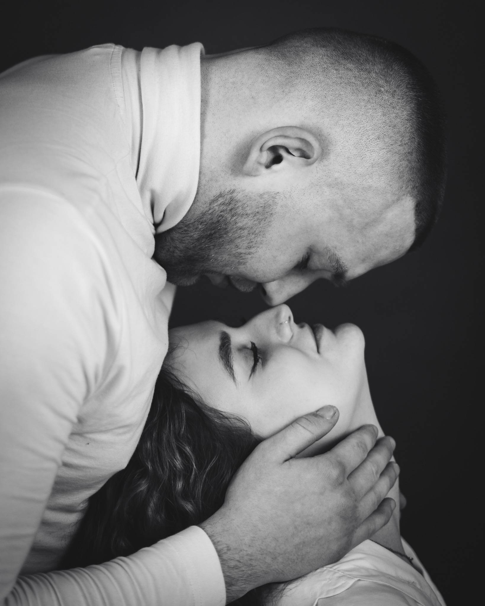 Czarno-białe zbliżenie pary: mężczyzna z krótkimi włosami obejmuje kobietę, delikatnie dotykając jej szyi dłonią; oboje mają zamknięte oczy.
