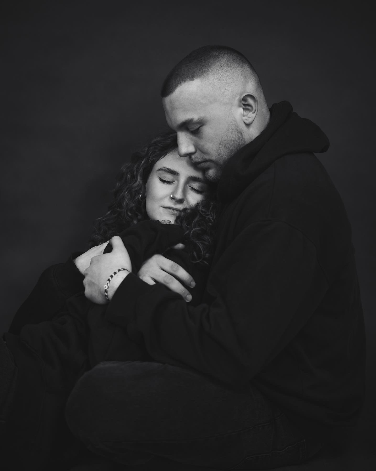 Czarno-białe zdjęcie objętej pary w bluzach z kapturami, kobieta z zamkniętymi oczami i nosem z kolczykiem, mężczyzna z krótkimi włosami przytula ją od tyłu, oboje siedzą na podłodze.
