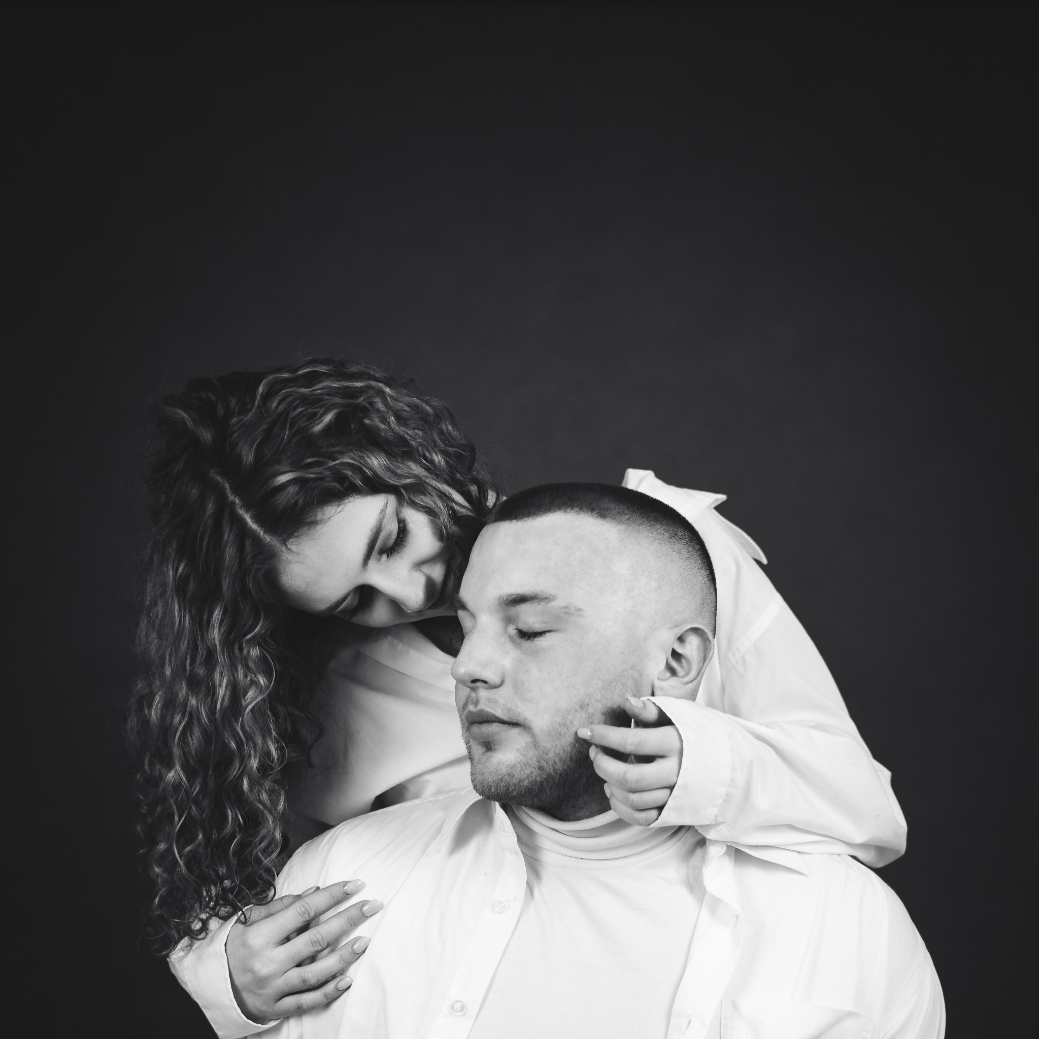 Czarno-białe portretowe ujęcie pary w białych koszulach na ciemnym tle; kobieta z kręconymi włosami obejmuje mężczyznę, dotykając jego policzka.