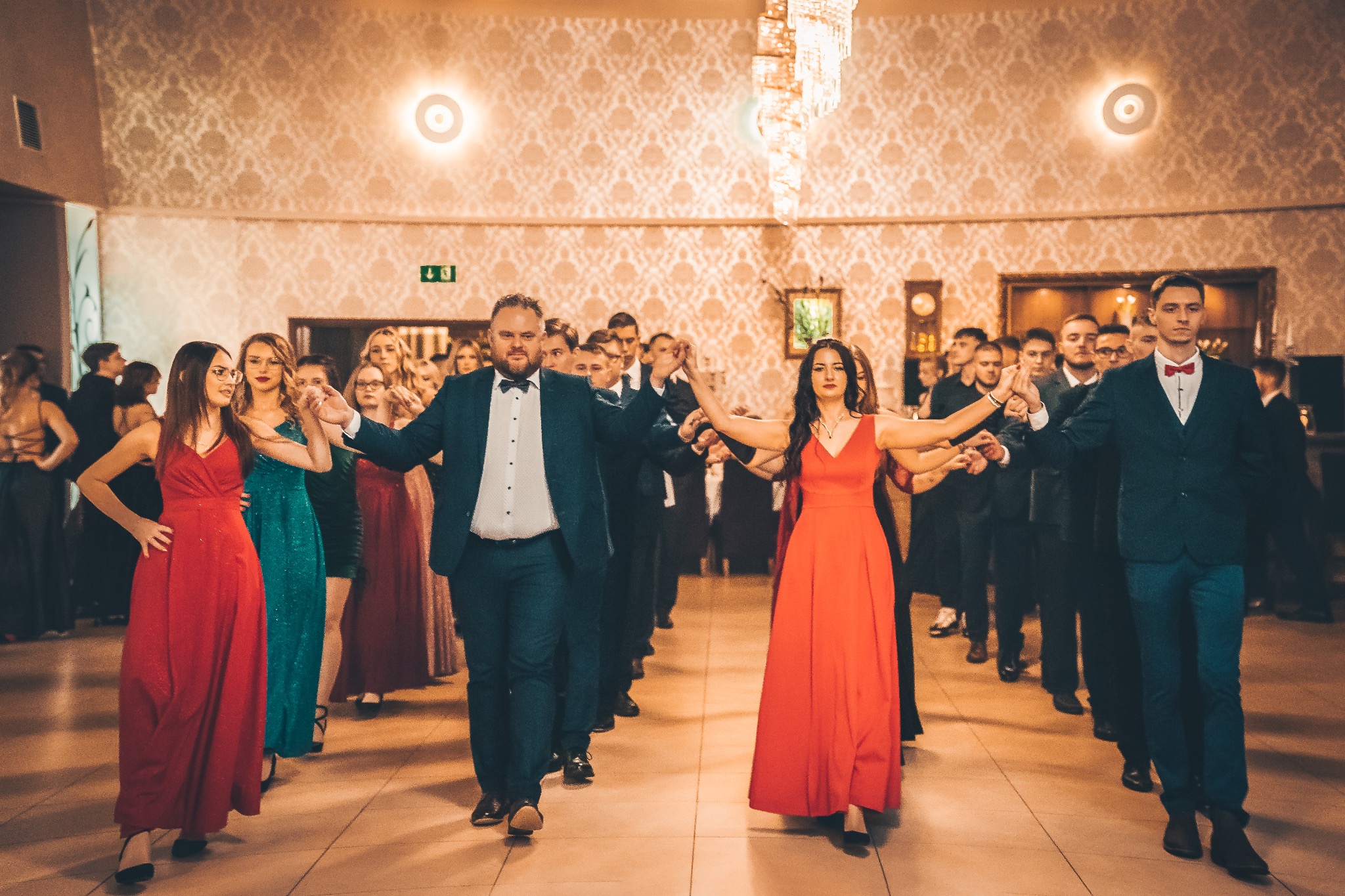 Grupa elegancko ubranych osób ustawiona w rzędach, tańcząca poloneza na sali balowej z żyrandolem i wzorzystą tapetą.