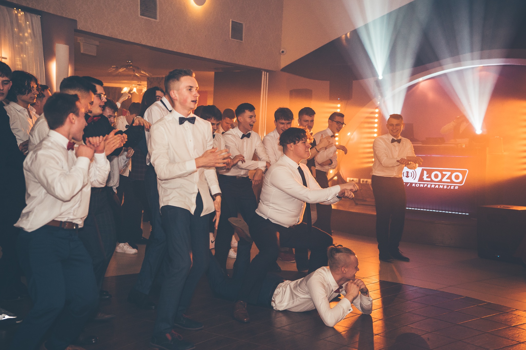 Grupa młodych mężczyzn w białych koszulach i muszkach tańczy na imprezie, oświetlona pomarańczowym światłem, z DJ-em w tle i jednym mężczyzną leżącym na parkiecie.