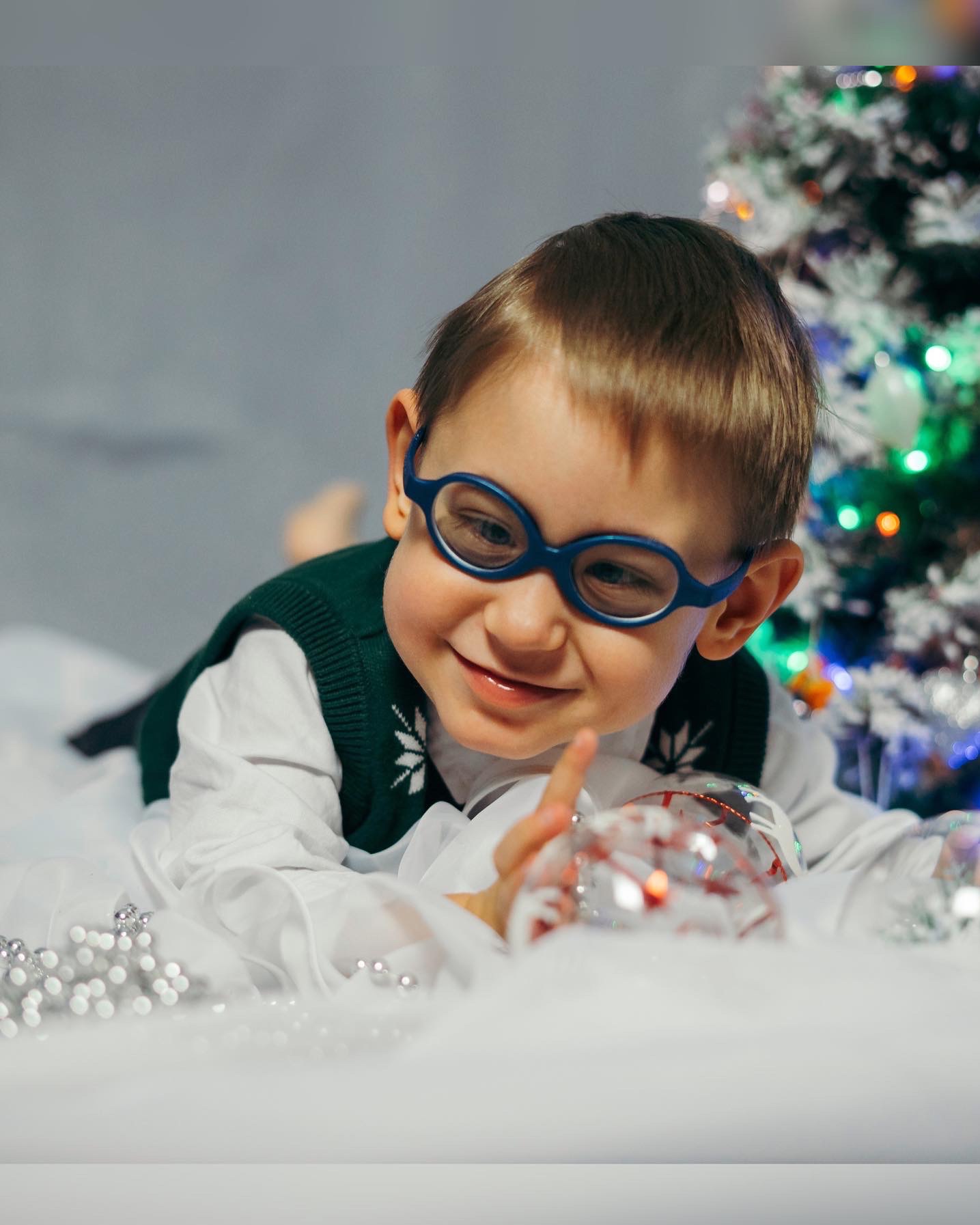 Uśmiechnięty chłopiec w okularach leży na białym tle z choinką w tle, trzymając przezroczystą bombkę z czerwonymi wzorami.