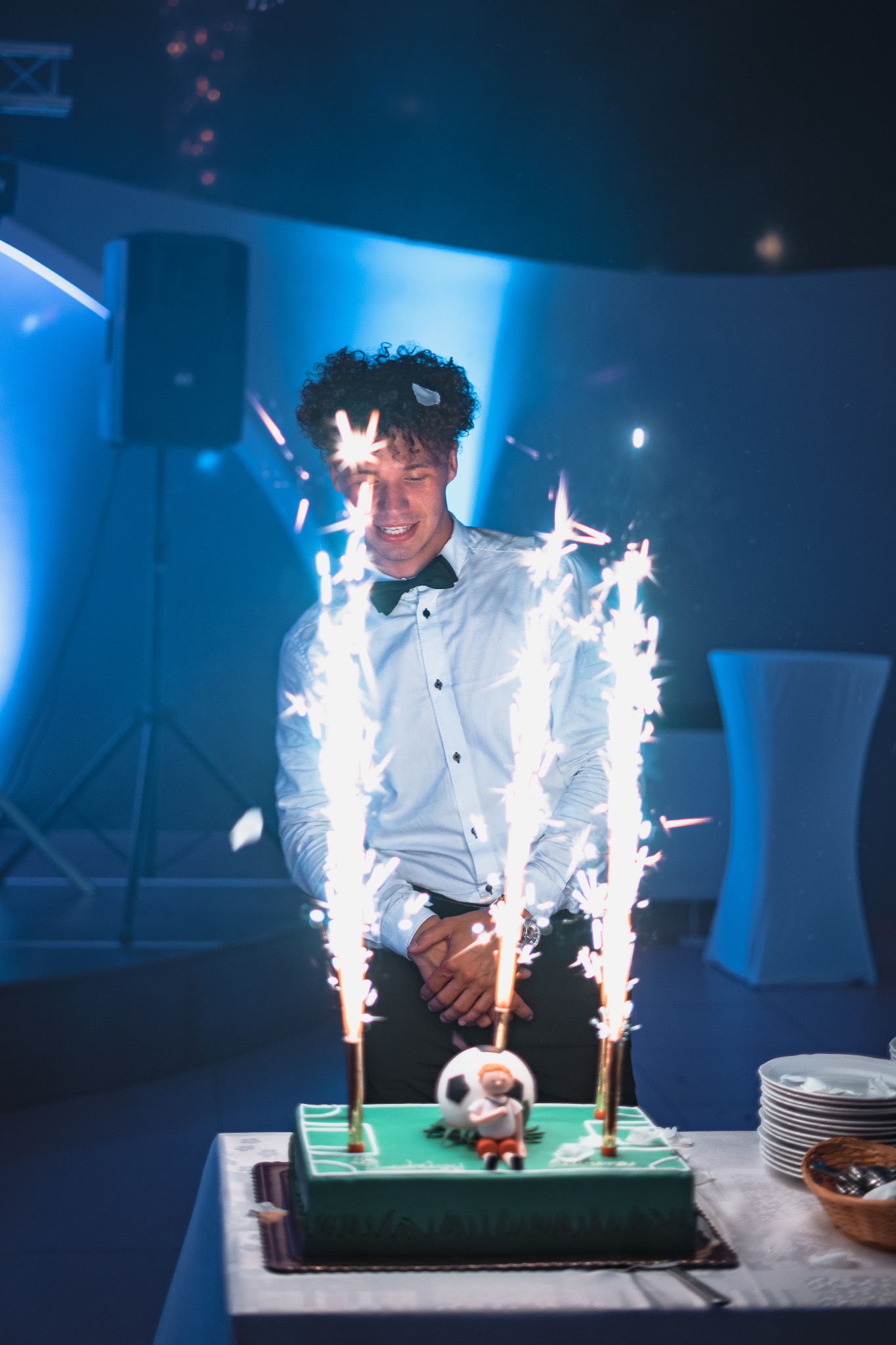 Uśmiechnięty mężczyzna w muszce stoi za tortem w kształcie boiska piłkarskiego, z fontannami iskier, w tle kolumna głośnikowa i dekoracje w niebieskim oświetleniu.