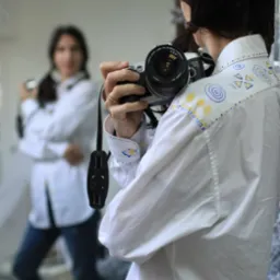 Kobieta fotografująca się w lustrze, ubrana w białą koszulę z ręcznie malowanymi, geometrycznymi wzorami na ramionach i mankietach, trzymająca aparat Canon EOS.