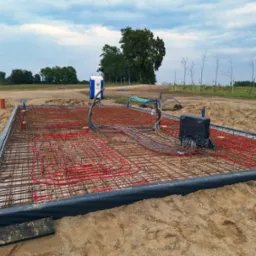 Ułożona instalacja ogrzewania podłogowego z czerwonymi rurami na zbrojeniu, przygotowana do wylewki. Widoczne obrzeża i otaczający teren budowy z toaletą przenośną.