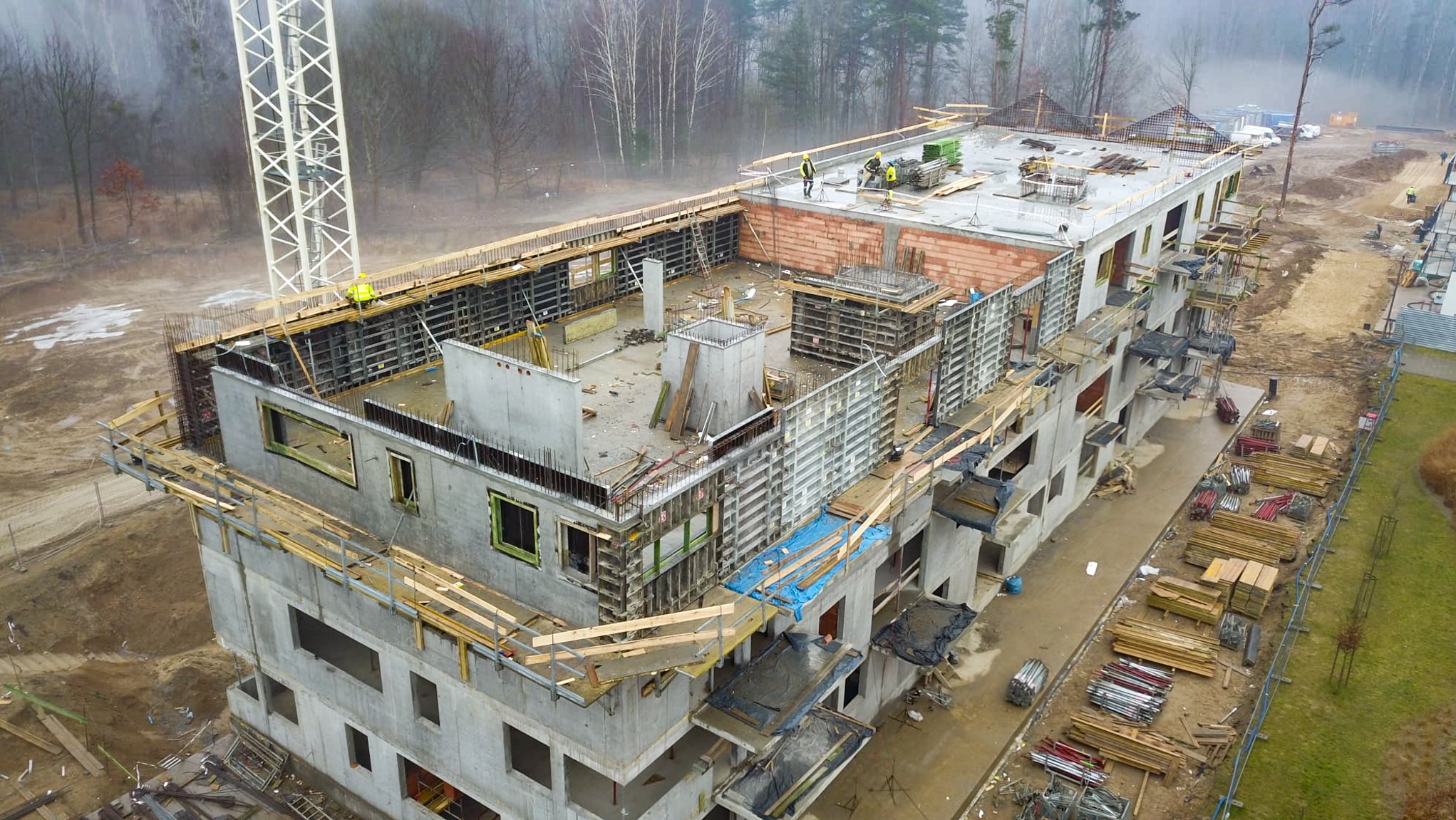 Wysoka perspektywa na budowę wielopiętrowego budynku żelbetowego w trakcie realizacji. Widoczne szalunki i robotnicy na dachu, teren budowy i drzewa w tle.