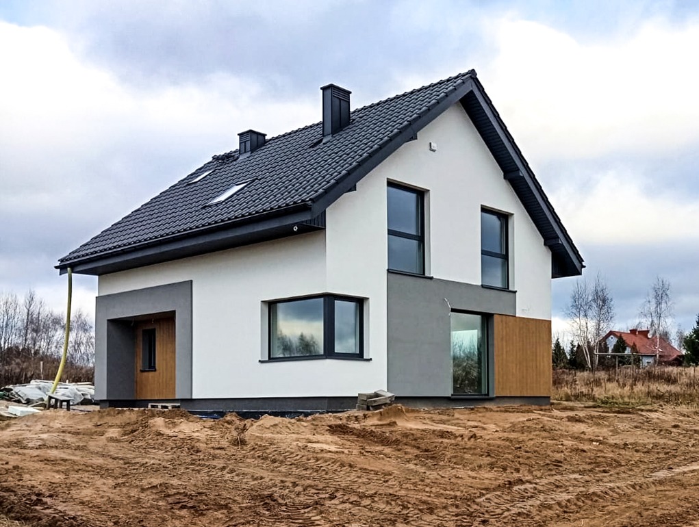Nowoczesny dom jednorodzinny z ciemną dachówką i grafitowymi oknami, częściowo wykończony drewnianą elewacją, na działce w trakcie zagospodarowania.
