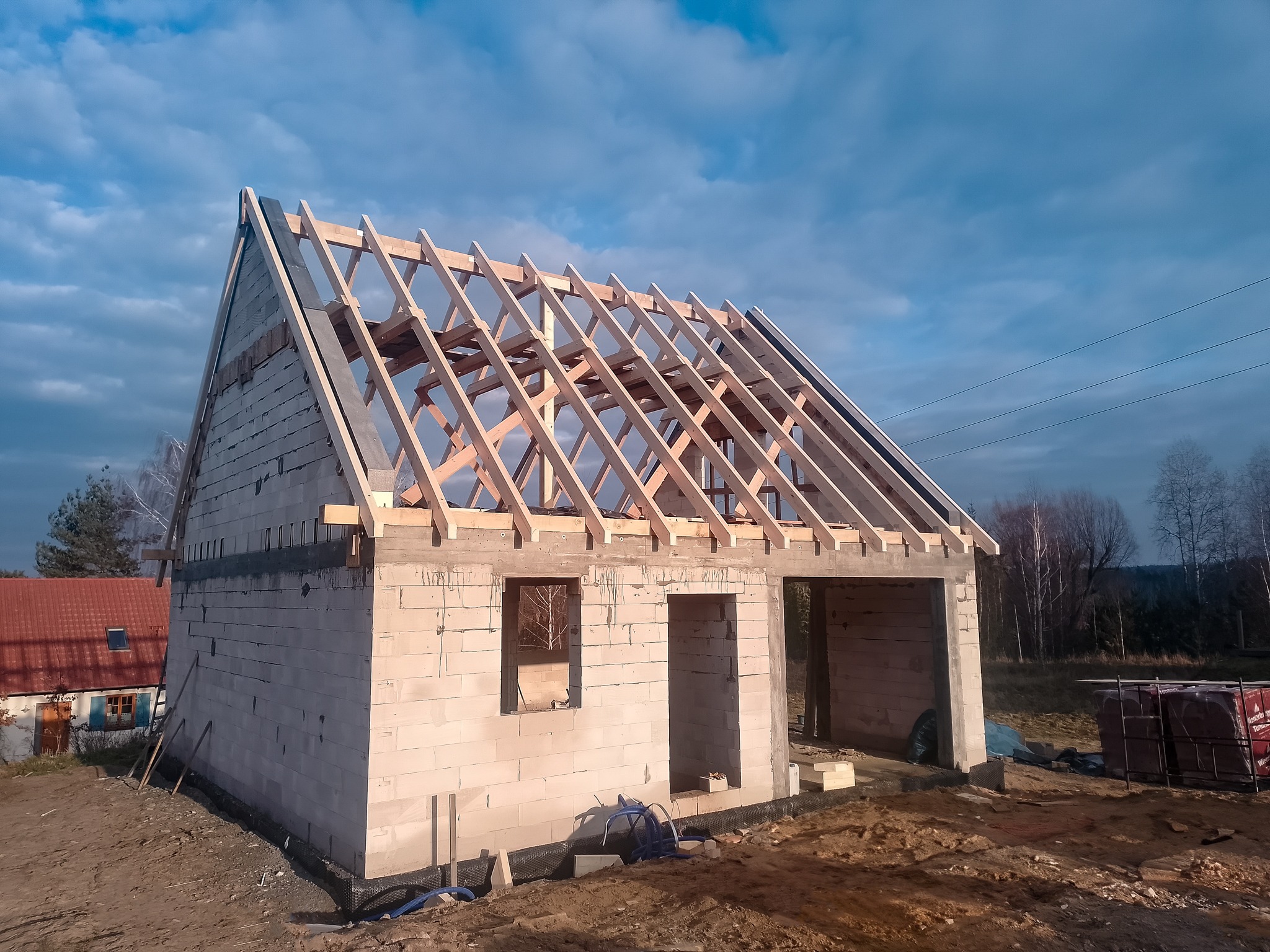 Budowa domu jednorodzinnego w trakcie realizacji: surowy stan z pustaków, widoczna konstrukcja dachu z drewna na tle błękitnego nieba.