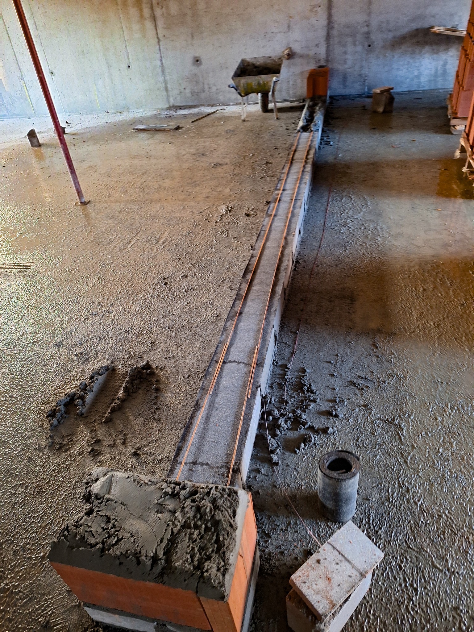 Wnętrze w trakcie murowania ściany z cegieł, widoczne mokre podłoże i elementy konstrukcyjne. Cegły połączone zaprawą, wyznaczona linia muru.