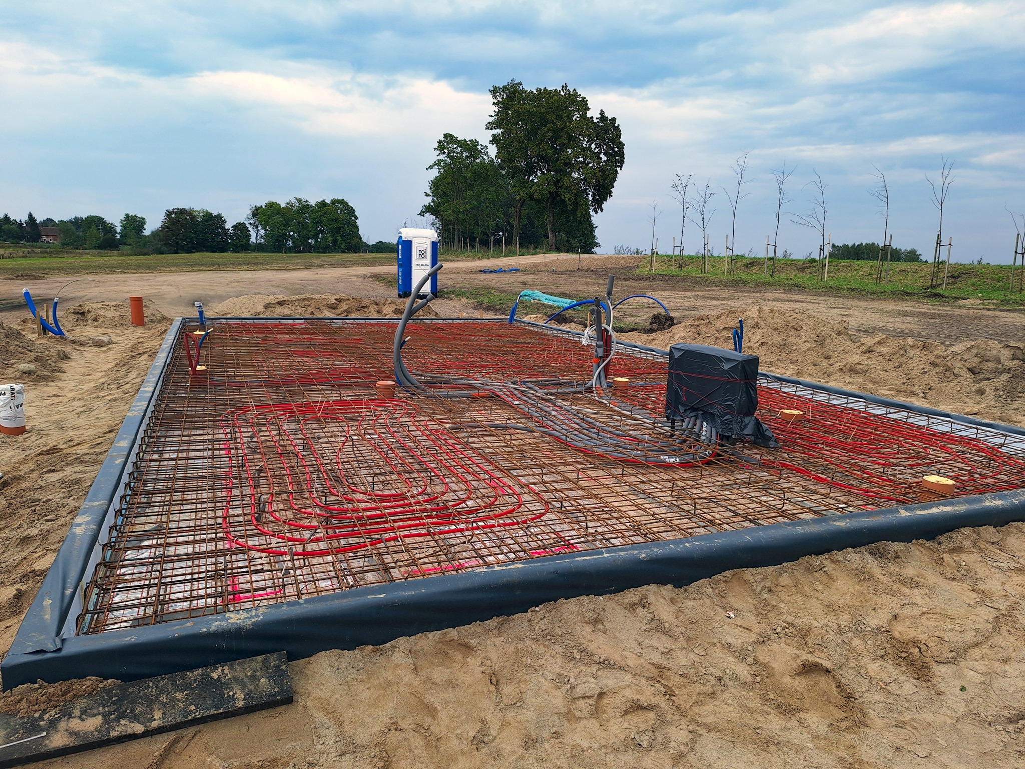 Ułożona instalacja ogrzewania podłogowego z czerwonymi rurami na zbrojeniu, przygotowana do wylewki. Widoczne obrzeża i otaczający teren budowy z toaletą przenośną.
