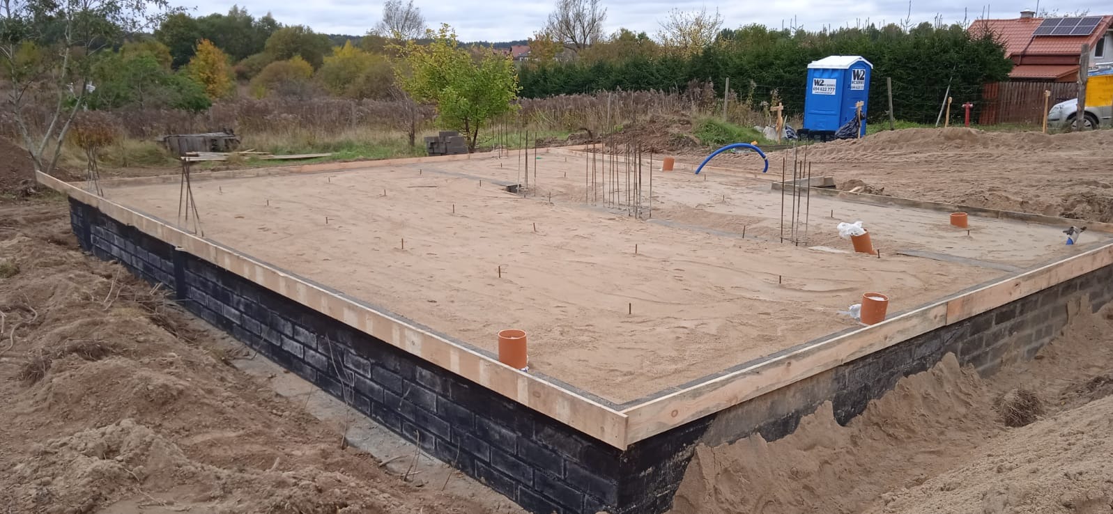 Fundamenty domu jednorodzinnego z izolacją, przygotowane do dalszych prac. Widoczne zbrojenia, szalunki i przyłącza kanalizacyjne. W tle TOI TOI i częściowo widoczny dom.