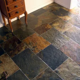 Ułożona pod kątem podłoga z wielobarwnego łupka afrykańskiego, oświetlona ciepłym światłem, z fragmentem drewnianej komody i złotego lustra w tle.