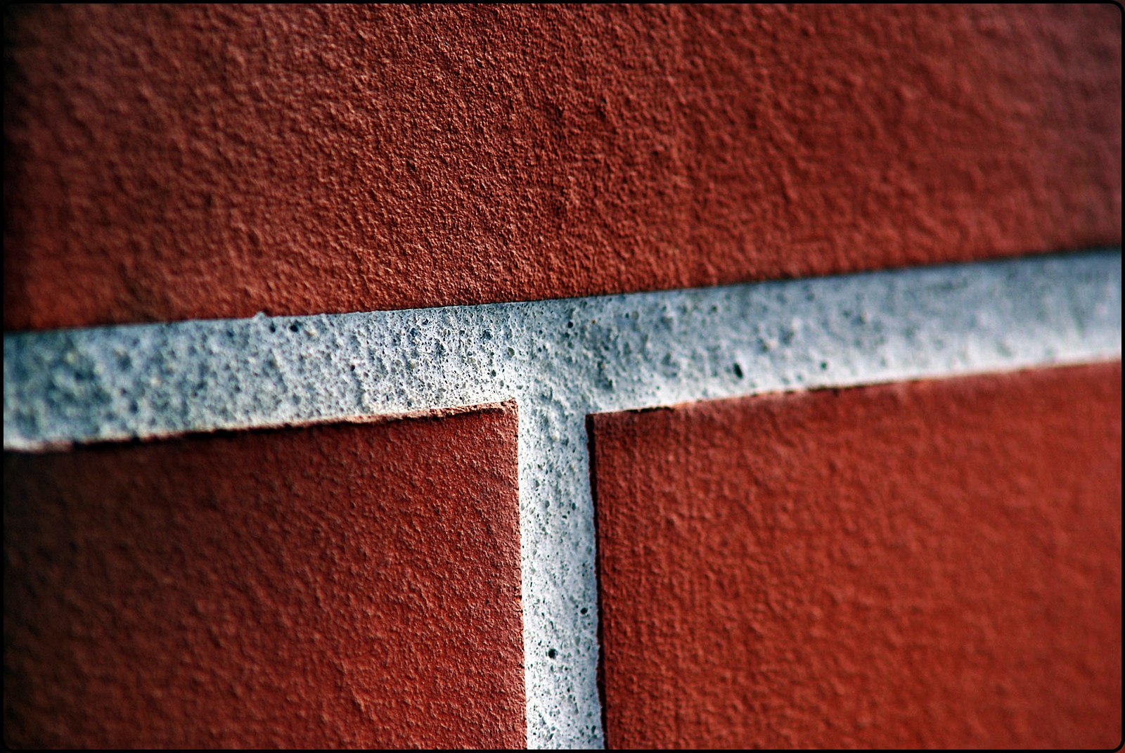 Zbliżenie na fragment ściany z czerwonego klinkieru z jasnoszarą spoiną tworzącą kształt litery T.