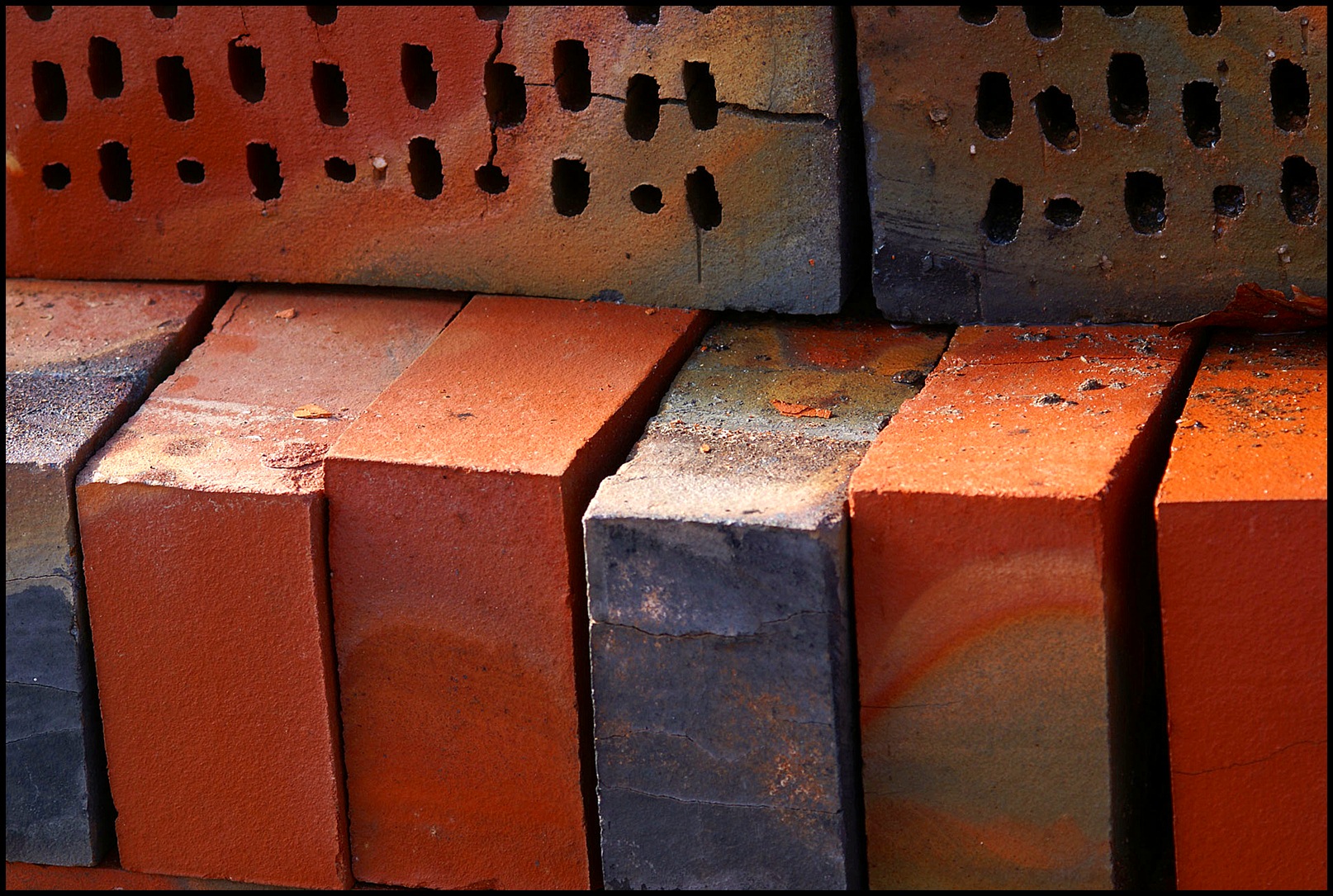 Cegły klinkierowe ułożone w stos, widoczne różne odcienie czerwieni i szarości, niektóre z otworami, z bliska, z drobnymi pęknięciami i śladami zabrudzeń.