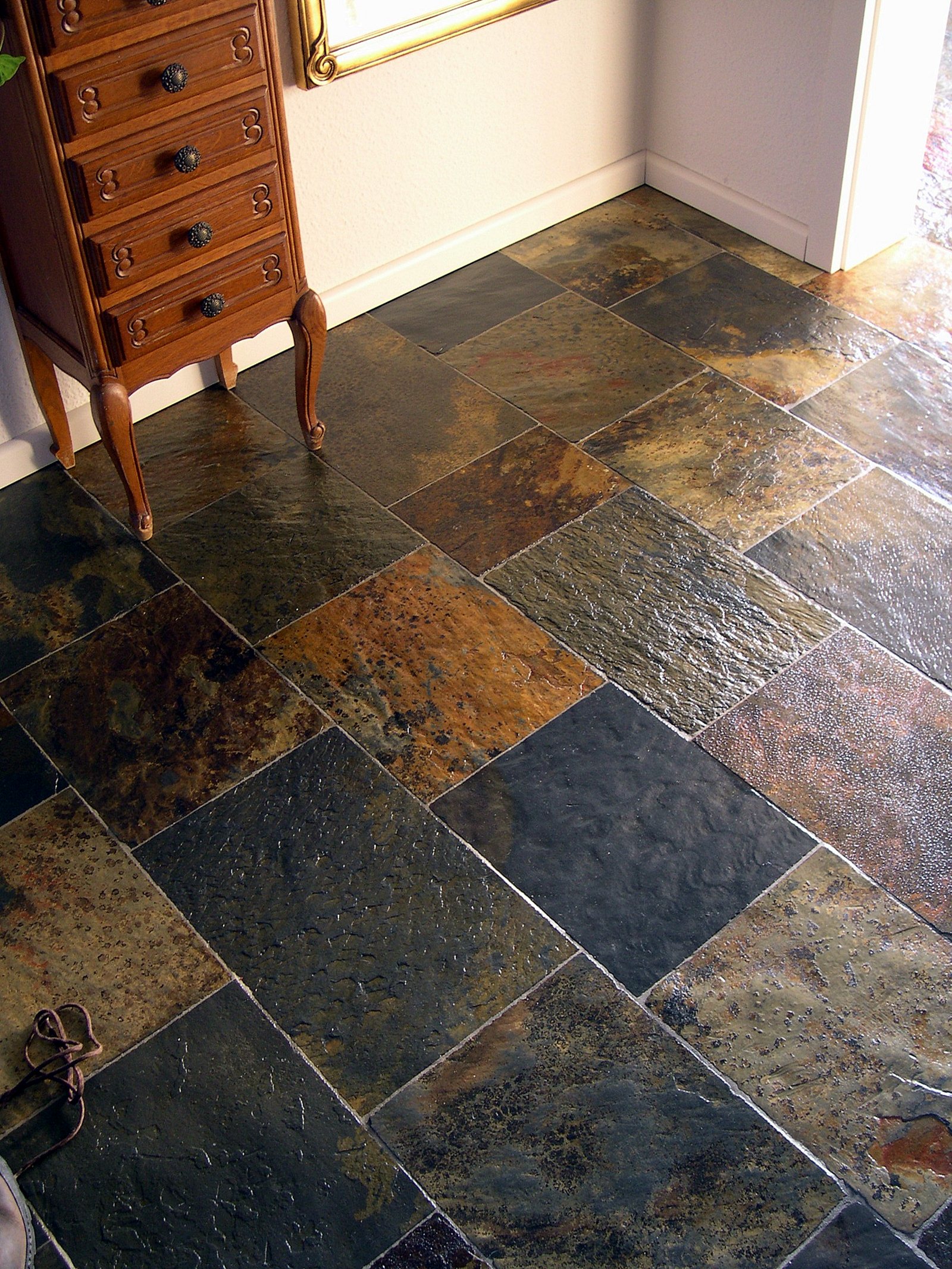 Ułożona pod kątem podłoga z wielobarwnego łupka afrykańskiego, oświetlona ciepłym światłem, z fragmentem drewnianej komody i złotego lustra w tle.